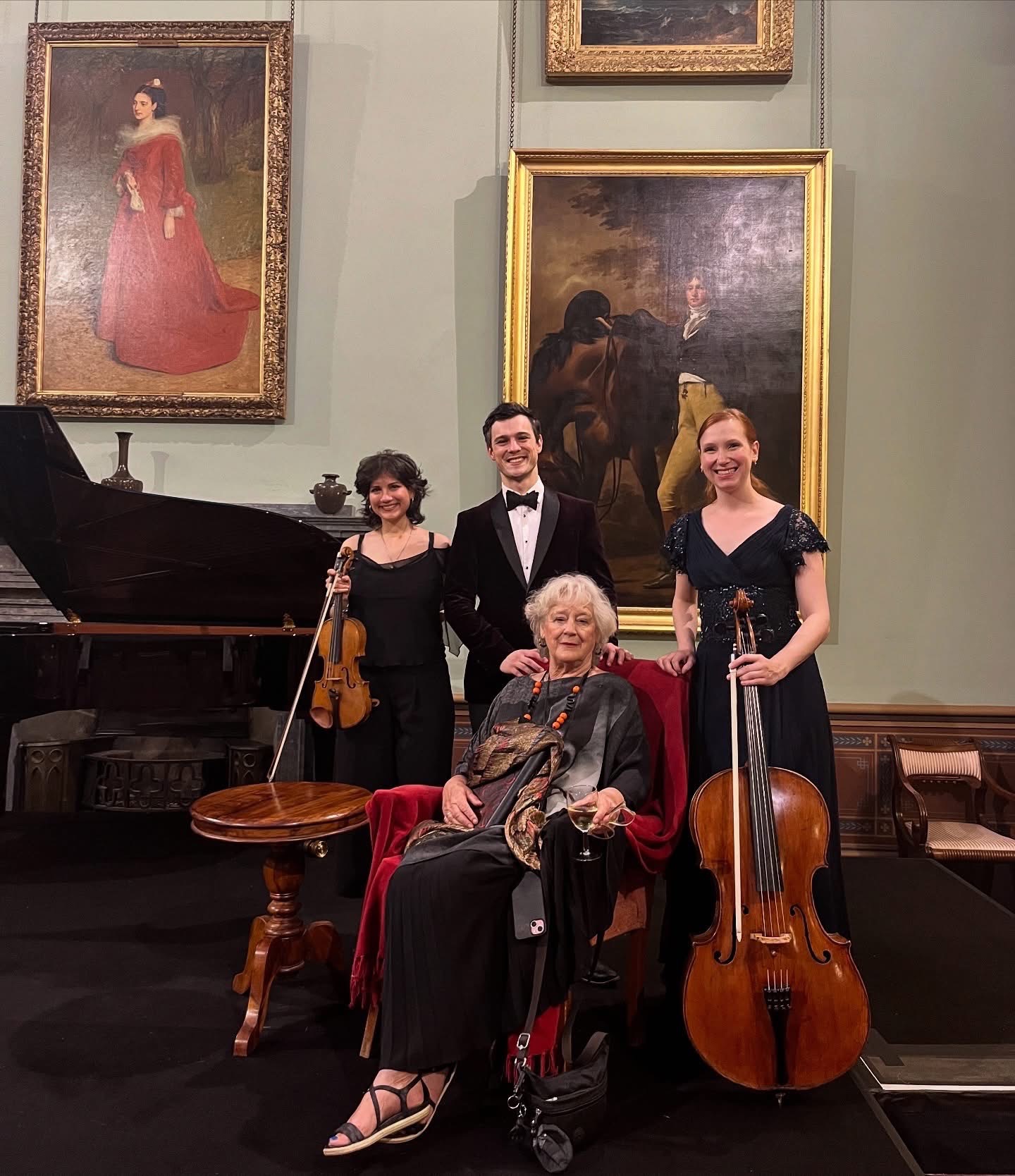 Three young musicians holding their instruments stand behind a seated older woman in a stately home.